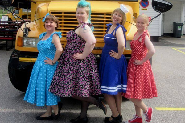 Neljä naista poseeraa 50-luvun mekoissa vanhan keltaisen bussin edessä. 